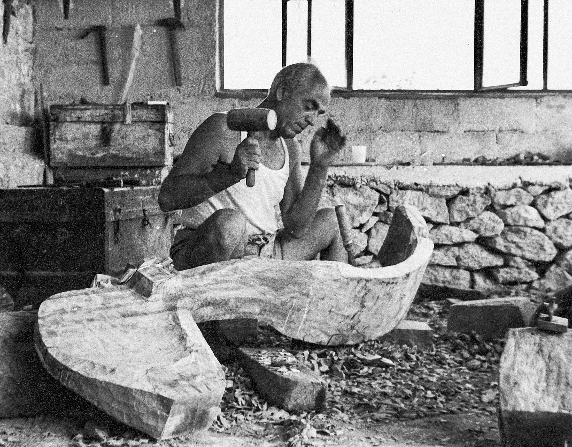 Το σπίτι-εργαστήριο του Χρήστου Καπράλου στην περιοχή Πλακάκια στην Αίγινα, χ.χ. (1965;). Ο γλύπτης στο υπόστεγο εργαστήριο λαξεύει μια ξύλινη μορφή με ματρακά και καλέμι. © Εθνική Πινακοθήκη-Μουσείο Αλεξάνδρου Σούτσου, Αρχείο Χρήστου Καπράλου